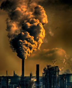 Sepia image showing a large plume of smoke travelling upwards from an industrial chimney in an indistinct view of a city environment.