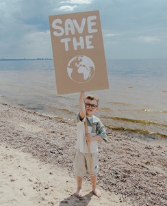 Image of a boy wearing long shorts and t-shirt, on the beach, with the sea in view. He's holding a placard reading 'Save the' with an image of a planet underneath.