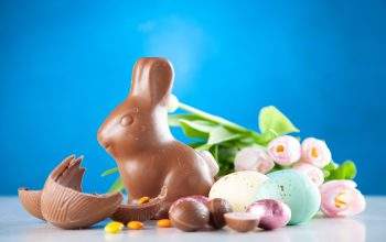 Chocolate rabbit next to a broken chocolate egg. Assorted painted eggs and a tulip flower in the background