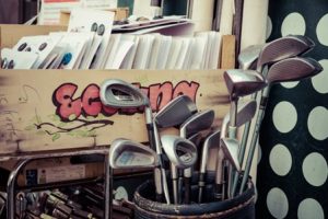 Image of golf clubs sticking out the top of a cylindrical container - with the club end on display. There is also a box full of buttons laid out on sheets of card.