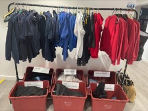 Photo of a clothes rail with hangers of children's school uniform - mostly polo tops and sweat shirts in colours black, blue, white, grey and red. Under the rail are labelled plastic boxes of other uniform items,. 