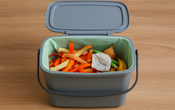 A silver grey food waste caddy on a kitchen counter top. The lid of the caddy is open revealing the caddy to be full with potato, onion and carrot peelings, and a used tea bag.