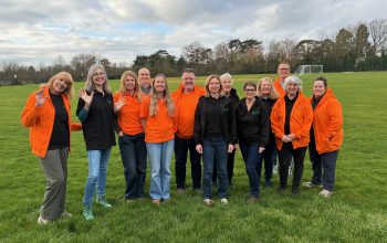 A group of 13 Environment Action Volunteers stood together in a green space wearing matching black and orange uniforms