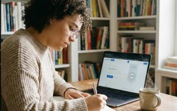 a female student sat at a desk in a book lined room using a laptop whilst writing in a naotepad.